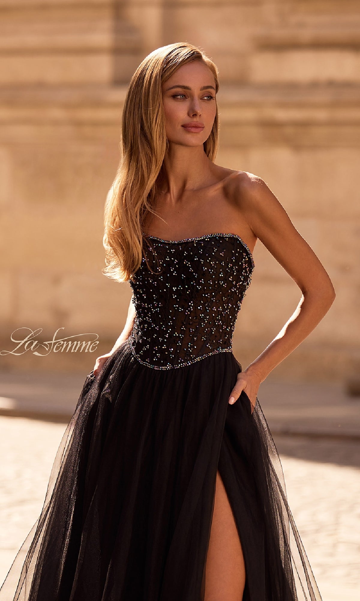 Woman wearing the Strapless Long Black Prom Ball Gown: La Femme 32856, featuring a beaded bodice and tulle skirt, standing outdoors with one hand in a pocket.